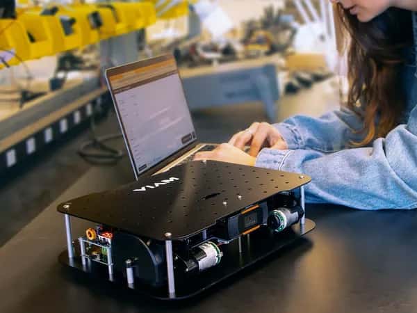 Viam Rover 2.0 — женщина сидит за ноутбуком и программирует. Перед ней — робот из двух ярусов чёрных прямоугольников с камерами и электроникой посередине.