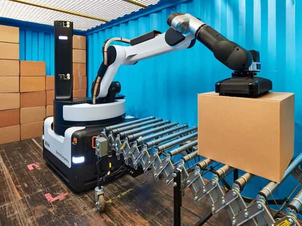 Робот Stretch от Boston Dynamics: крупная чёрно-серая мобильная база с шарнирным манипулятором и вакуумным захватом, снимающим картонную коробку с конвейера. На заднем плане — стопка коробок.