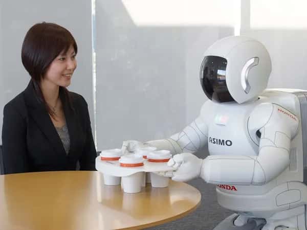 Женщина сидит за столом, пока робот Asimo подносит подставку с четырьмя чашками кофе.