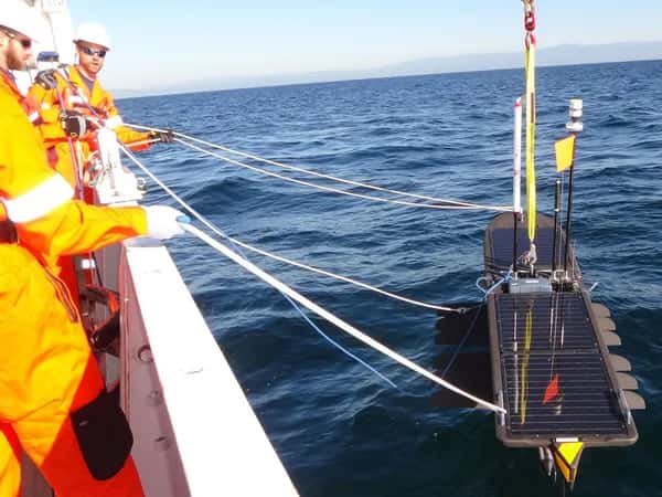 Wave Glider — рабочие в касках и оранжевых комбинезонах стоят на борту судна, удерживая белые тросы, прикреплённые к длинному роботу, похожему на доску: он покрыт солнечными панелями, оснащён несколькими мачтами и находится на воде.