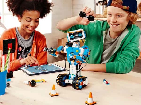 Lego Boost  — двое подростков используют планшет для программирования робота из кубиков Lego: гуманоид с колёсными гусеницами вместо ног, двумя руками и милым лицом с голубыми глазами и оранжевыми бровями.