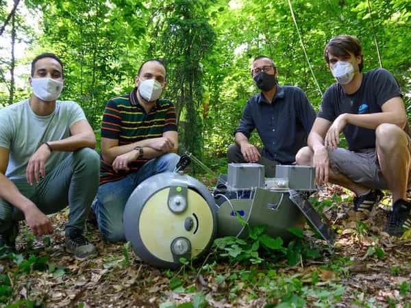 SlothBot — четыре человека в масках сидят на корточках позади большого робота-ленивца.