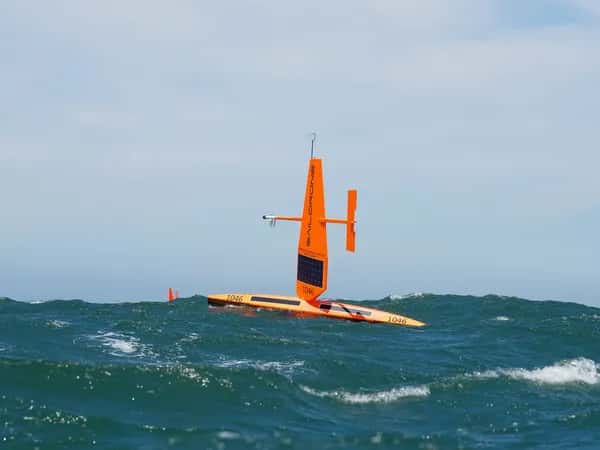 Saildrone — оранжевое беспилотное морское судно с горизонтальным корпусом на воде, большим парусом с солнечными панелями и рулём, параллельным морю.