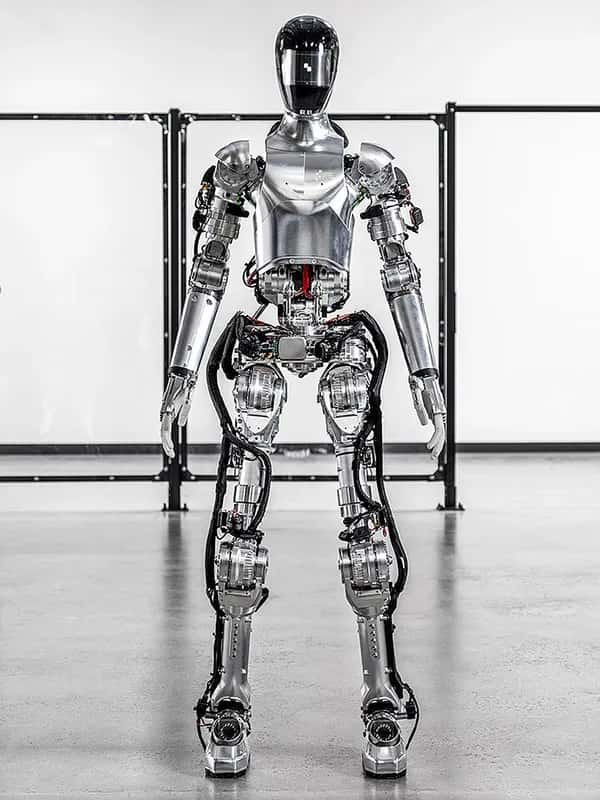 Робот Figure 01 — блестящий серебристый двуногий гуманоид с руками и шлемообразной головой — стоит на складе.