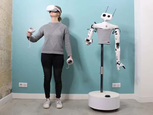 Reachy 2 — женщина в VR-очках с предметами в руках стоит рядом с гуманоидным торсом на стойке и мобильной платформе, который повторяет её позу.