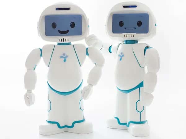 QTrobot — два дружелюбных маленьких гуманоида позируют вместе. У них простые белые корпуса, руки и округлая лицевая панель с экраном, на котором отображается улыбающаяся мультяшная рожица.
