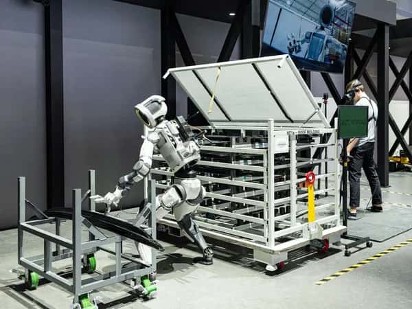 Atlas — гуманоидный робот перемещает детали автомобиля. На заднем плане — человек в VR-очках.