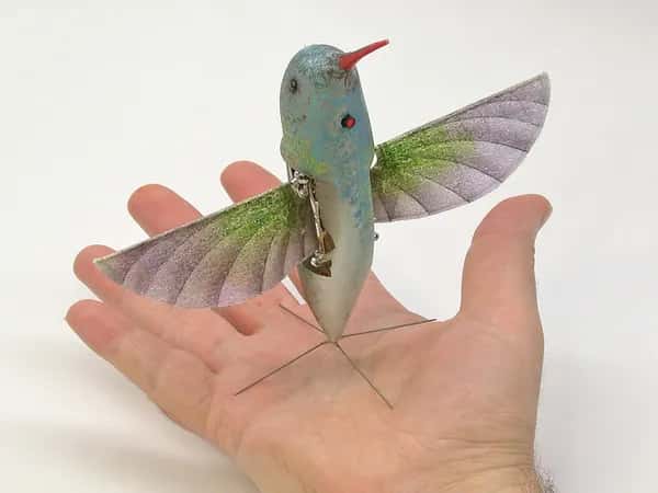 Nano Hummingbird — робот, имитирующий колибри: крылья, длинный тонкий оранжевый клюв — стоит на четырёхштырьковом основании на ладони человека.