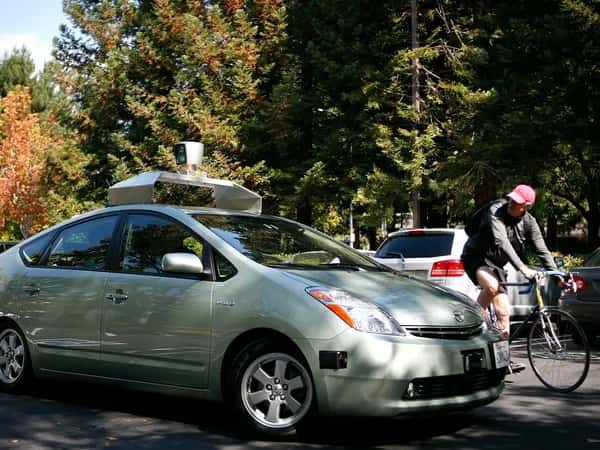 Google Self-Driving Car — человек на велосипеде поворачивается посмотреть на автономную машину с лидарами и другим оборудованием.