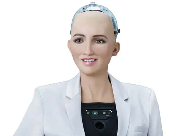 Sophia — робот с женской внешностью: реалистичная кожа персикового оттенка, зелёные глаза, тонкий нос и розовые губы с лёгкой улыбкой. Верхняя часть головы — прозрачный кожух с электроникой. Робот одет в белый пиджак поверх механического торса с чёрной панелью, оснащённой камерами и датчиками.
