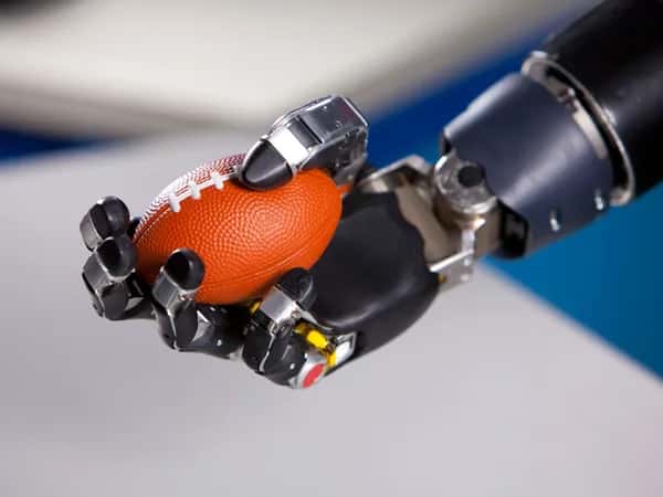 Modular Prosthetic Limb — чёрная роботизированная рука с пятью суставчатыми пальцами, имитирующая человеческую, держит оранжевый мини-мяч для американского футбола.