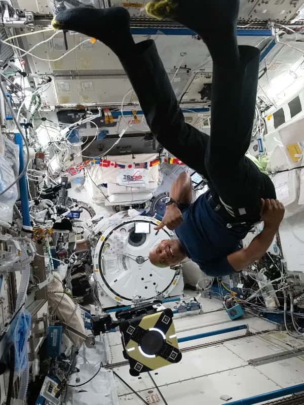 Улыбающийся астронавт вверх ногами парит рядом с роботом Astrobee на космической станции, окружённой электроникой.