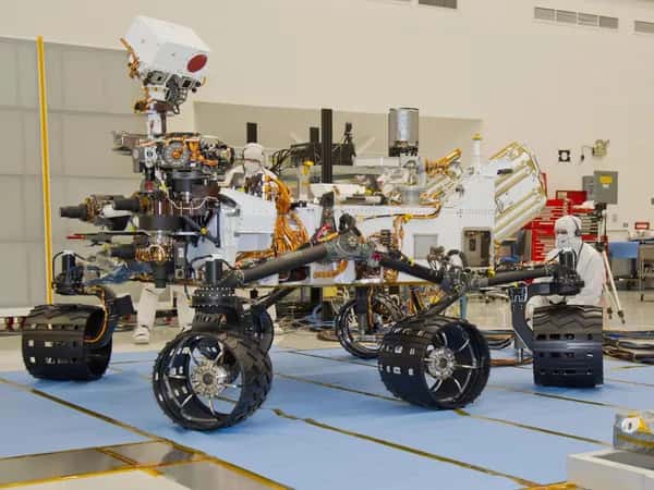 Curiosity — человек в защитном костюме чистой комнаты стоит на коленях рядом с большим шестиколёсным роботизированным аппаратом, оснащённым камерами и приборами.