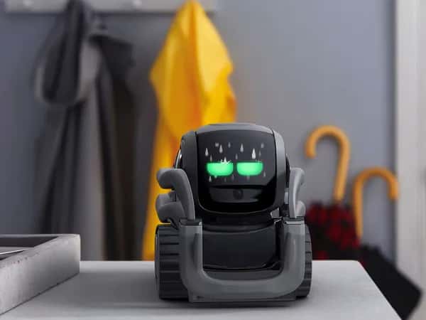Vector 2.0 — vector — простой компактный чёрный колёсный робот размером меньше ладони с двумя светящимися зелёными глазами, стоит на поверхности в доме.