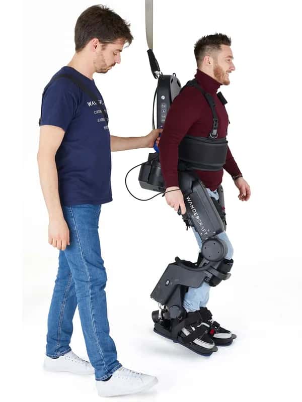 Atalante X — улыбающийся мужчина делает шаг в экзоскелете Wandercraft: ремни охватывают его ноги и торс. Рядом стоит второй человек, держащий страховочный ремень.