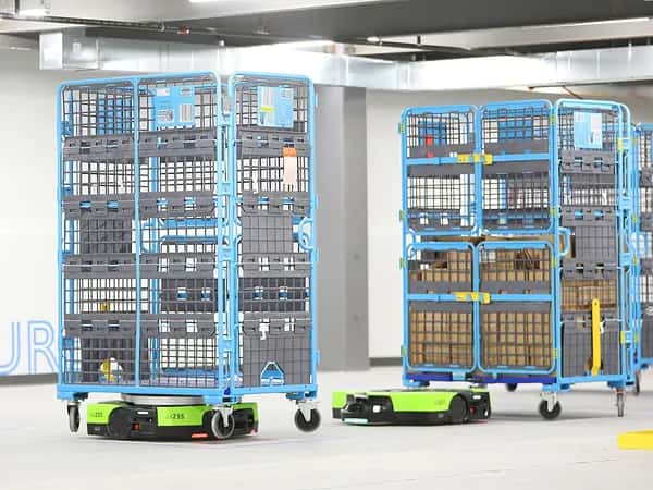 Proteus — два низкорамных зелёных мобильных робота перевозят по складу синие многоярусные стеллажи.