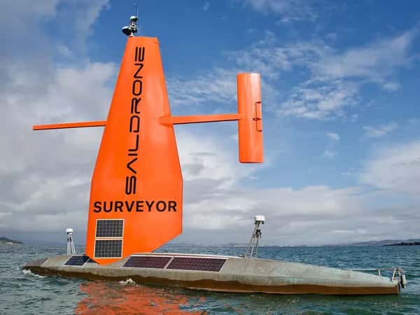 Saildrone — беспилотное надводное судно. Горизонтальный серебристый корпус с солнечными панелями, вертикальный оранжевый парус с солнечными панелями и руль, параллельный поверхности воды.