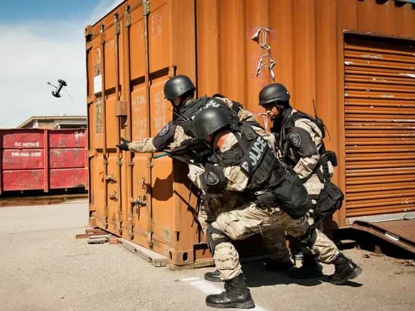 Трое бойцов SWAT в снаряжении и камуфляже укрываются за стеной, один бросает Throwbot.
