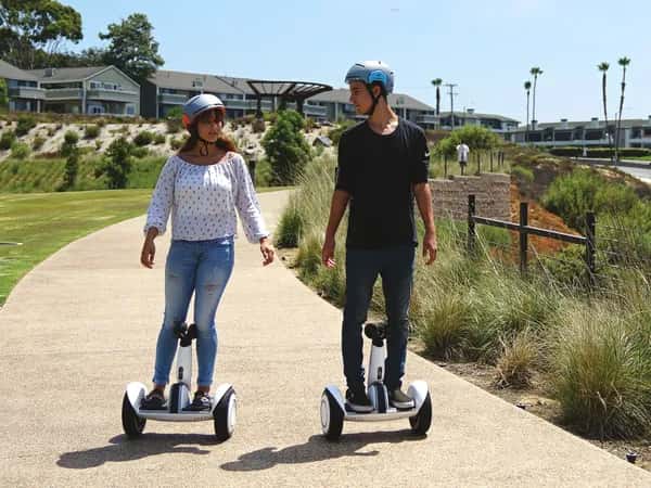 Segway — двое в шлемах едут на мини-сигвеях: платформа на двух колёсах и вертикальная стойка между ног водителя.