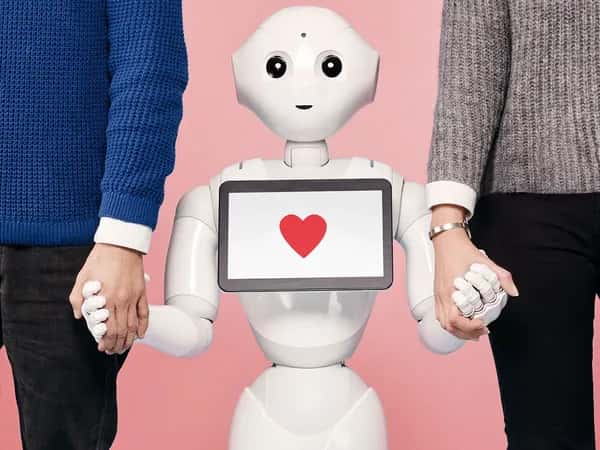Pepper — дружелюбный белый робот с сердечком на планшете держит за руки двух взрослых людей.