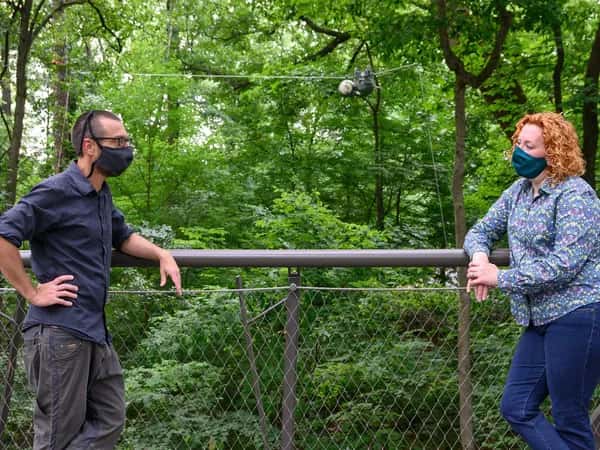 Двое в масках облокотились на забор на улице. Вдали за ними робот SlothBot висит на тросе.