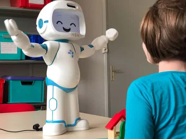 QTrobot — робот стоит на столе с раскинутыми руками и смеющимся выражением лица. Рядом смотрит на него ребёнок.
