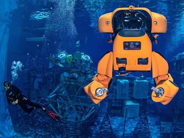 Aquanaut — оранжевый полугуманоидный робот-субмарина с захватными руками под водой рядом с аквалангистом.