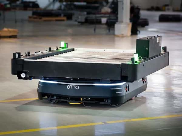 OTTO — приземистый квадратный чёрно-серебристый мобильный робот перевозит крупный объект по заводскому цеху.