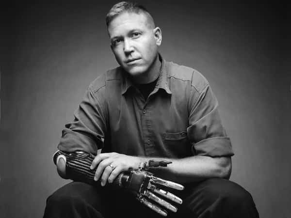 Modular Prosthetic Limb — чёрно-белая современная фотография: сидящий мужчина держит свою роботизированную руку живой рукой.
