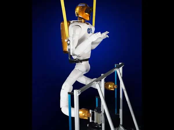 Robonaut 2 — робот подвешен в страховочной системе. К корпусу прикреплены две белые ноги с мягкими суставами и золотистыми захватами, которыми он манипулирует объектами снизу.
