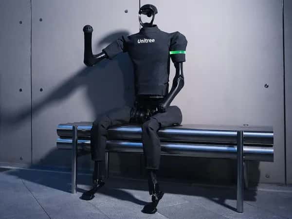 Unitree H1 — серый двуногий гуманоид в футболке Unitree сидит на скамейке с поднятой рукой.