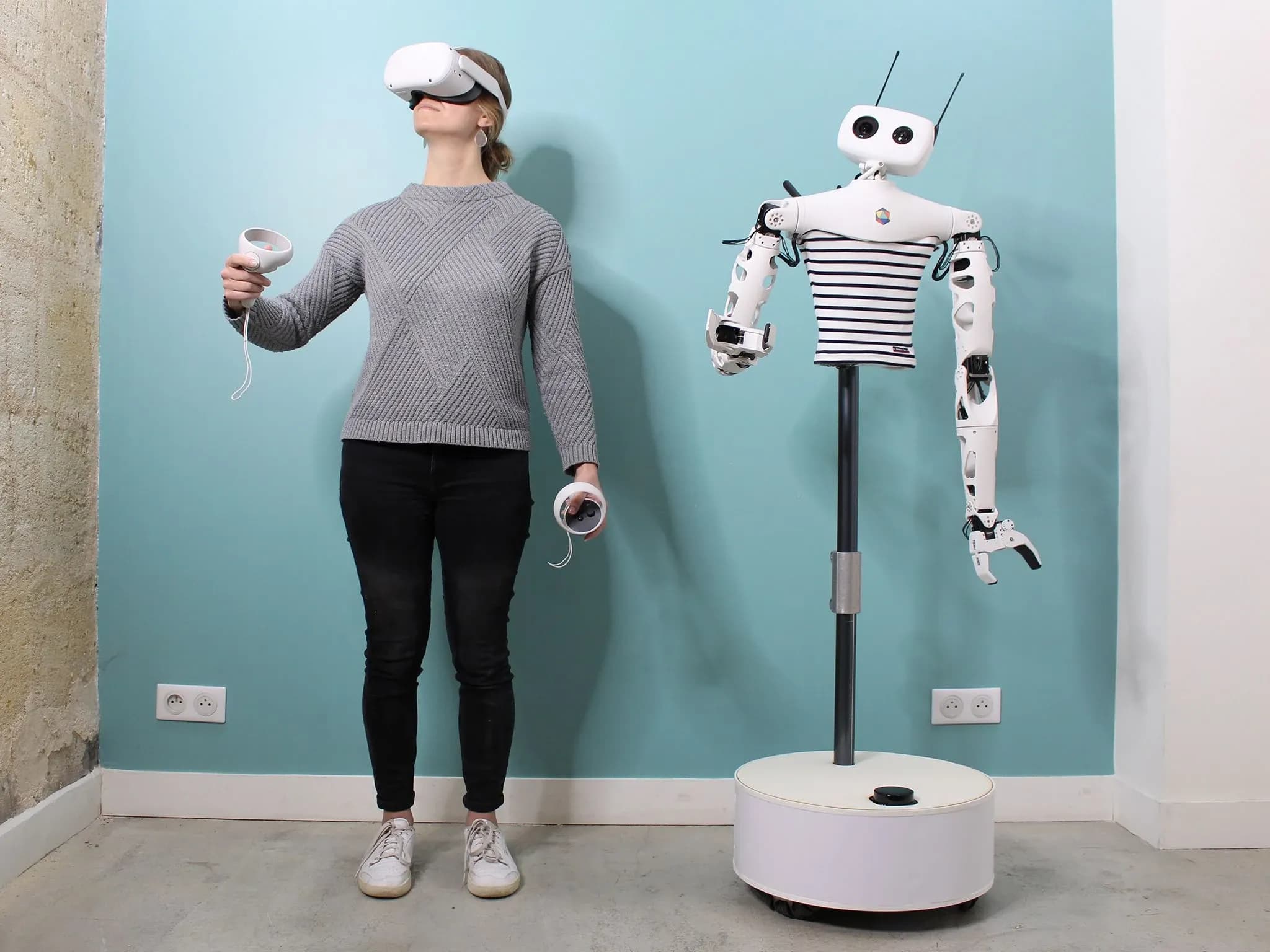 Женщина в VR-очках с предметами в руках стоит рядом с гуманоидным торсом на стойке и мобильной платформе, который повторяет её позу.