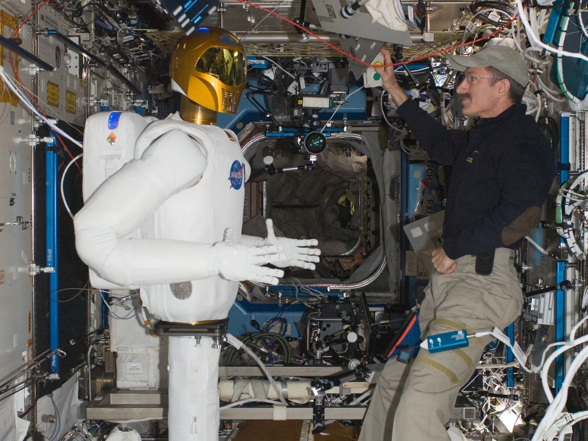 Robonaut и астронавт в обычной одежде находятся внутри космической станции, заполненной оборудованием.