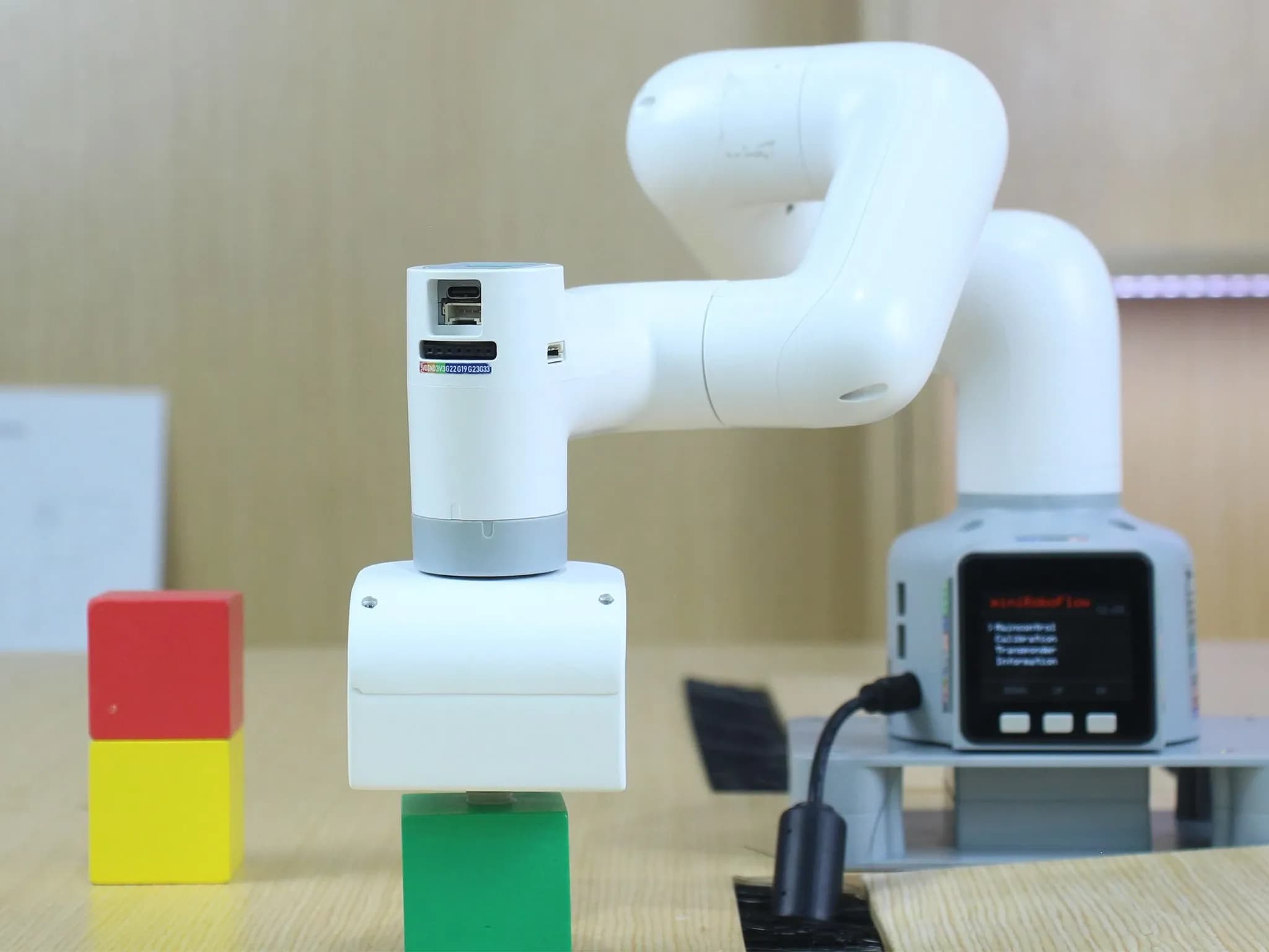 Белый робот-манипулятор myCobot 280 поднимает зелёный кубик со стола, на котором также стоят красный и жёлтый кубики.
